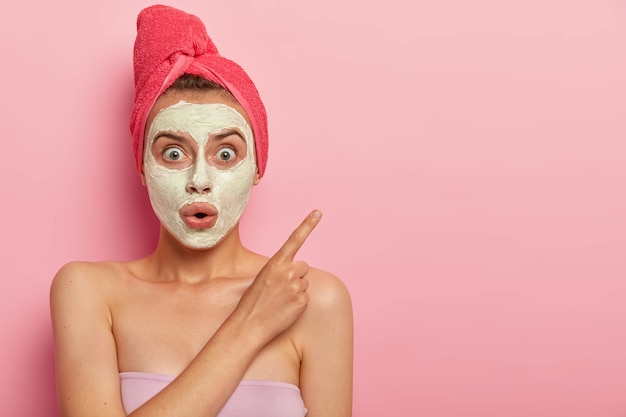 Foto van verrast sprakeloze vrouw draagt gezichtsmasker, heeft schoonheidsprocedures thuis, geschokte uitdrukking, wijst opzij met wijsvinger, handdoek gewikkeld op nat haar