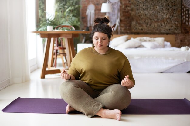 Foto van vreedzame kalme jonge mollige vrouw blootsvoets zittend op yoga mat thuis, mudra gebaar maken, mediteren met gesloten ogen. Evenwicht, meditatie, harmonie, zen en wellness-concept