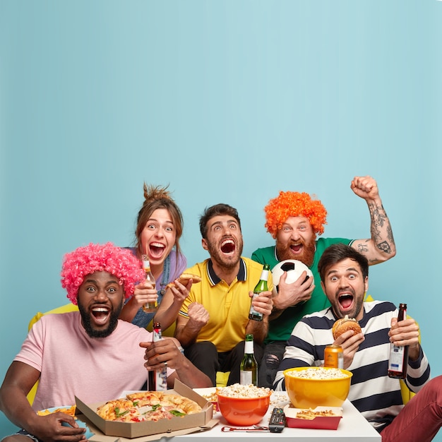 Foto van vrienden kijken naar voetbalwedstrijd, vieren het doel, balken vuisten, kijken naar sportwedstrijden, hebben een heerlijke snack, drinken koud bier, brengen vrije tijd thuis door. Tijdverdrijf en vermaak