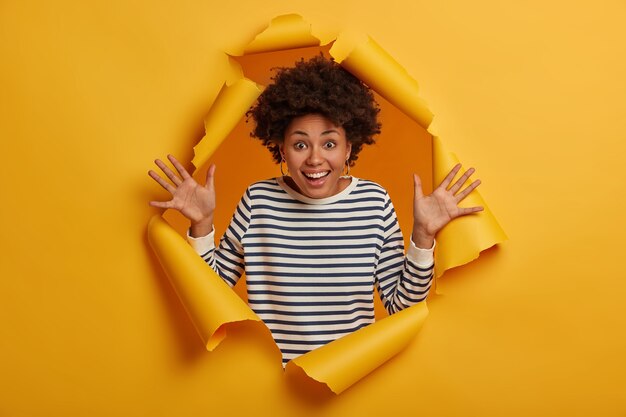 Foto van vrolijke gekrulde vrouw werpt palmen, verheugt zich op laatste ontmoeting met goede vriend, gekleed in gestreepte casual trui, gebaren van geluk, poses in gescheurde gescheurde papier achtergrond