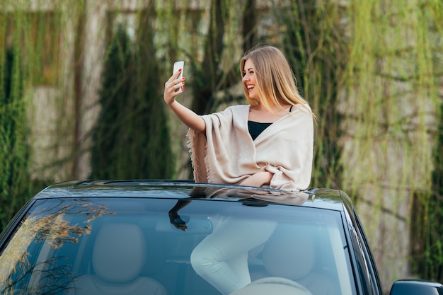 Foto van vrolijke jonge vrouw die zonnebril en opgeheven handen op het zonnedak van de luxeauto draagt