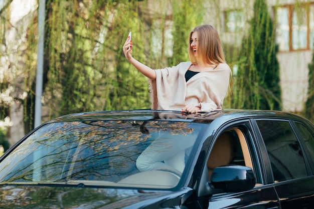 Foto van vrolijke jonge vrouw die zonnebril en opgeheven handen op het zonnedak van de luxeauto draagt