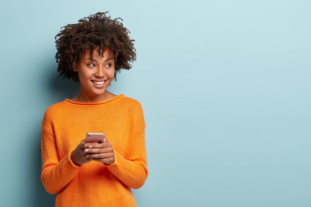 Foto van vrolijke opgetogen Afro-Amerikaanse vrouw typen sms op moderne mobiele telefoonapparaat, geniet van een goede internetverbinding