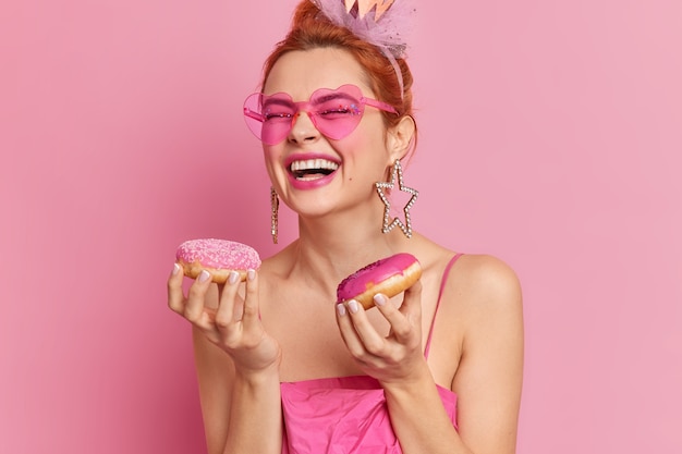 Foto van vrolijke stijlvolle roodharige vrouw in trendy zonnebril glimlacht in het algemeen houdt smakelijke donuts heeft zoetekauw