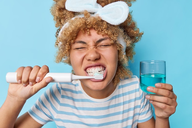 Foto van vrouw met krullend haar reinigt tanden met elektrische tandenborstel ondergaat ochtend mondhygiëne houdt glas mondwater terloops gekleed geïsoleerd over blauwe achtergrond stomatologie concept