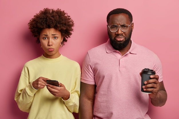 Gratis foto foto van wanhopig nors afro-amerikaans echtpaar leest slecht nieuws in sociale netwerken, drinkt afhaalkoffie, trieste vrouw wijst op display van smartphone