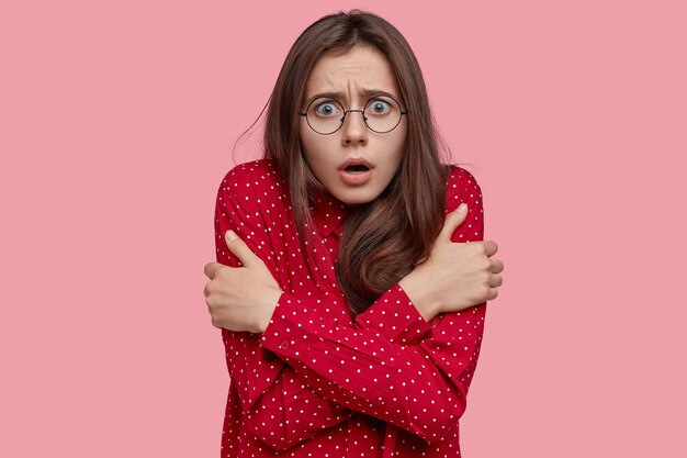 Foto van wanhopige vrouw steekt handen over de borst, voelt angst en opwinding, merkt angstaanjagende scène op, gekleed in een rode blouse met stippen