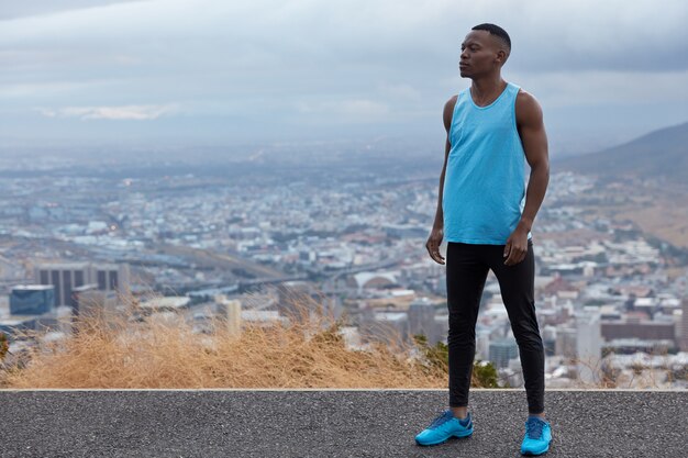 Foto van zwarte sportman draagt blues sneakers, vest en legging, modellen tegen hoogte boven de horizon, grote stad en bergen, vrije ruimte voor uw informatie. Panoramisch zicht