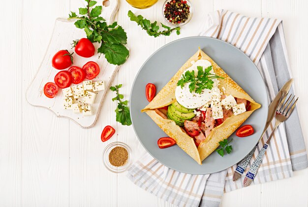 Franse keuken. Ontbijt, lunch, snacks. Pannenkoeken met gepocheerd ei, fetakaas, gebakken ham, avocado en tomaten op witte tafel. Bovenaanzicht