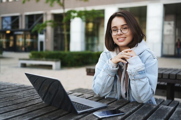 Freelance, mensen en onderwijsconcept. Vrolijk jong aantrekkelijk meisje zit alleen op een bankje, universiteit, werkt op afstand met laptop, mobiele telefoon, kijk weg met een tevreden glimlach.