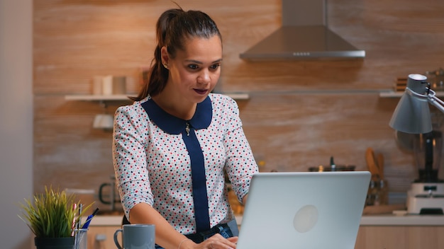 Freelancer ontvangt goed nieuws op laptop die vanuit huis in de keuken werkt. Enthousiaste gerichte werknemer die moderne technologienetwerken draadloos gebruikt om overuren te studeren, schrijven, zoeken