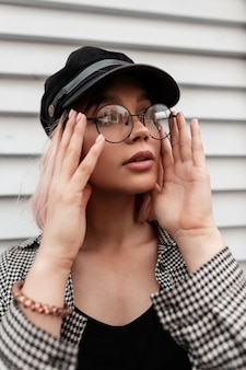 Fris portret van een mooie jonge tienervrouw zet een vintage bril op met een hoed in een mode casual geruit hemd in de buurt van een houten houten muur Premium Foto