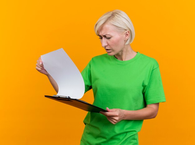 Fronsen blonde vrouw van middelbare leeftijd houden en kijken naar klembord geïsoleerd op gele muur