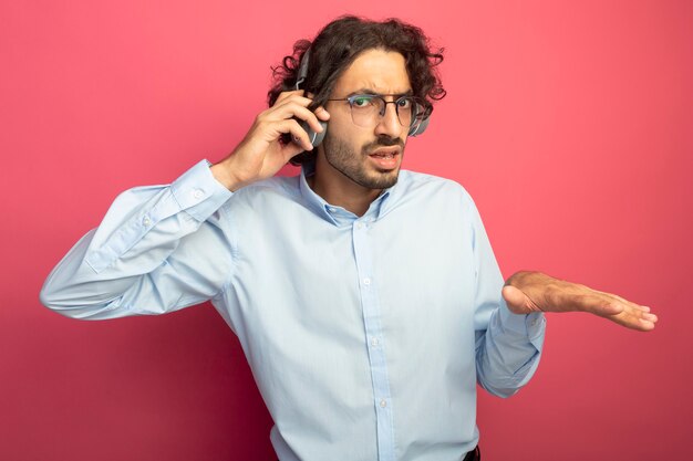 Fronsen jonge knappe man met bril en koptelefoon kijken naar voorzijde grijpen koptelefoon hand in de lucht houden uitleggen iets geïsoleerd op roze muur