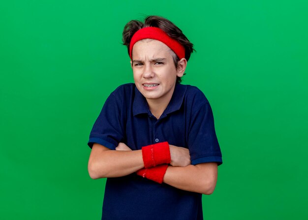 Fronsen jonge knappe sportieve jongen hoofdband en polsbandjes met beugels dragen permanent met gesloten houding kijken voorzijde geïsoleerd op groene muur met kopie ruimte