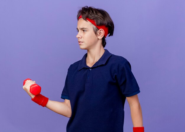 Fronsen jonge knappe sportieve jongen met hoofdband en polsbandjes met beugels kijken naar kant houden halter geïsoleerd op paarse muur