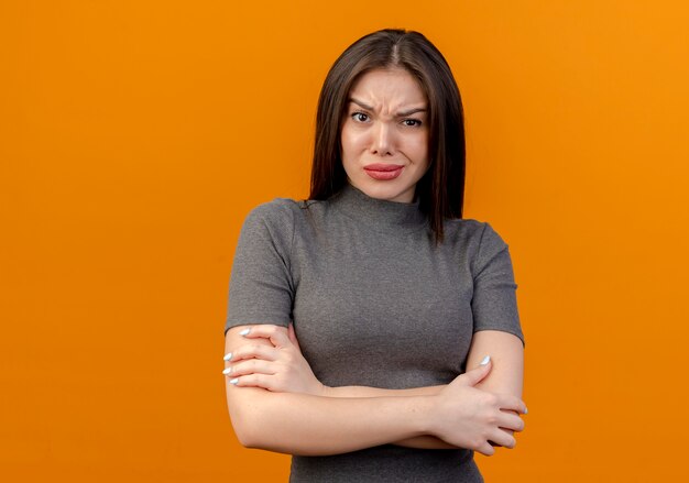 Fronsen jonge mooie vrouw met gesloten houding geïsoleerd op een oranje achtergrond met kopie ruimte