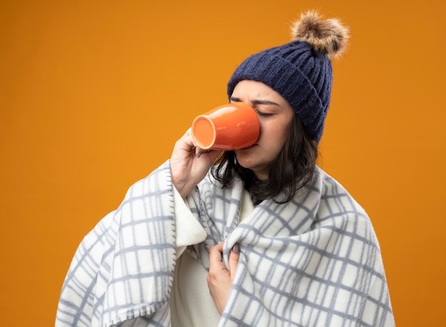 Fronsen jonge zieke vrouw met gewaad winter hoed verpakt in geruite kopje thee drinken grijpende plaid met gesloten ogen geïsoleerd op oranje muur