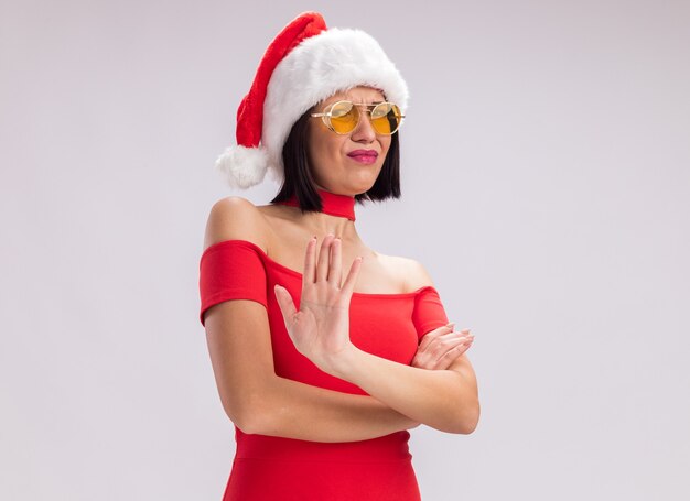 Fronsend jong meisje met een kerstmuts en een bril die naar de camera kijkt en een weigeringsgebaar doet geïsoleerd op een witte achtergrond met kopieerruimte