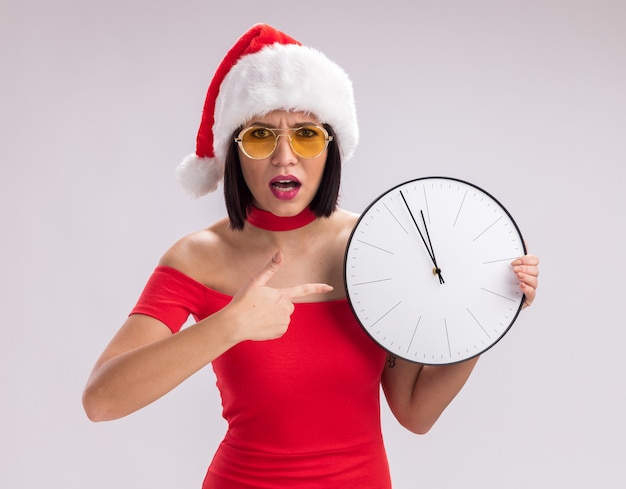 Fronsend jong meisje met een kerstmuts en een bril die naar de klok wijst en naar de camera kijkt die op een witte achtergrond wordt geïsoleerd
