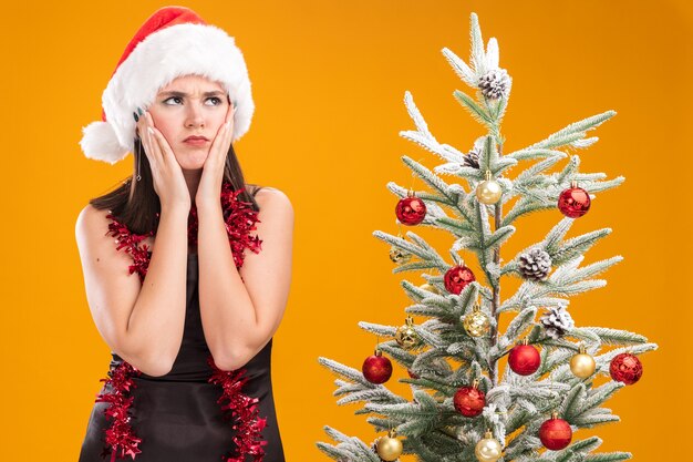 Fronsend jong mooi Kaukasisch meisje met een kerstmuts en een klatergoudslinger om de nek die in de buurt van een versierde kerstboom staat en de handen op het gezicht houdt en omhoog kijkt geïsoleerd op een oranje achtergrond