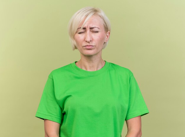 Fronsende blonde Slavische vrouw van middelbare leeftijd die zich met gesloten ogen bevindt die op olijfgroene achtergrond wordt geïsoleerd