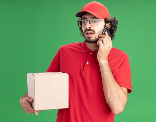 Fronsende jonge blanke bezorger in rood uniform en pet met een bril die een kartonnen doos vasthoudt die aan de telefoon praat en neerkijkt op een groene muur