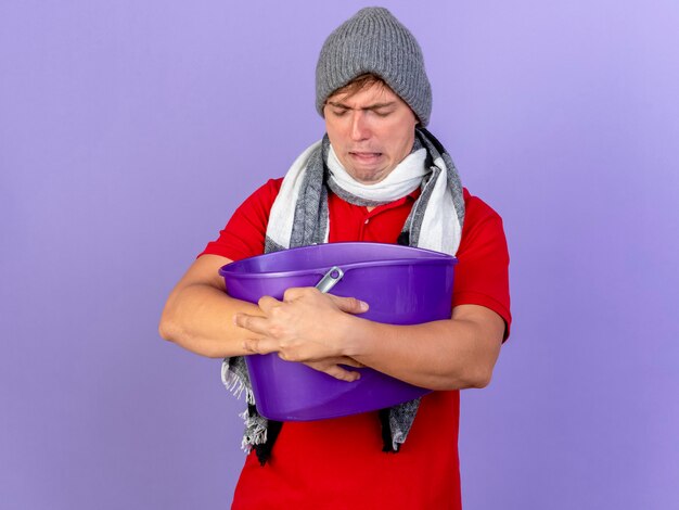 Fronsende jonge knappe blonde zieke man met muts en sjaal met plastic emmer met gesloten ogen met misselijkheid geïsoleerd op paarse muur met kopie ruimte