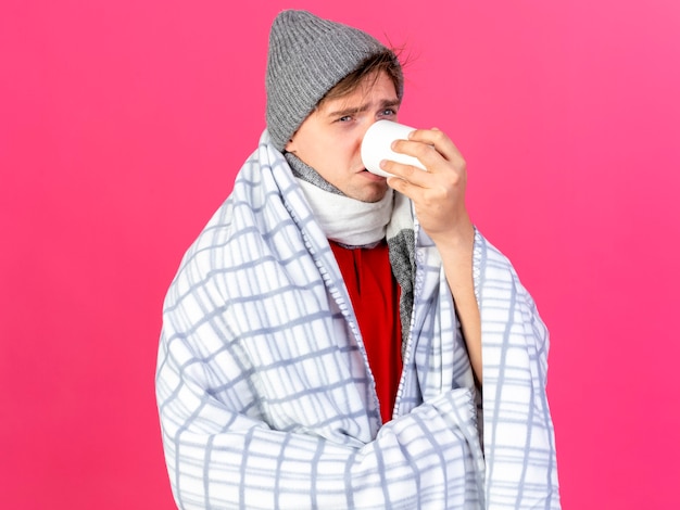 Fronsende jonge knappe blonde zieke man met winter muts en sjaal gewikkeld in een plaid op zoek recht drinkend kopje thee geïsoleerd op roze muur