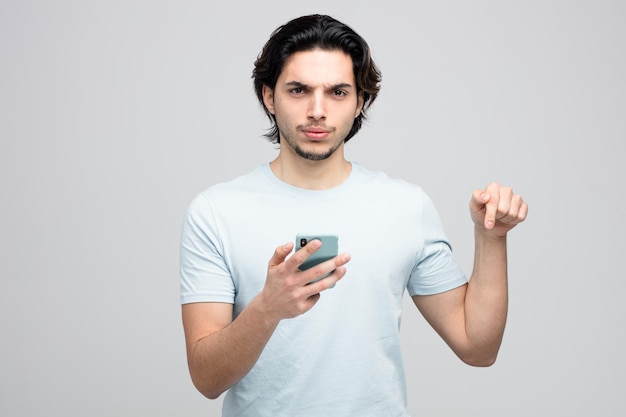 Fronsende jonge knappe man met mobiele telefoon kijkend naar camera die naar beneden wijst geïsoleerd op een witte achtergrond