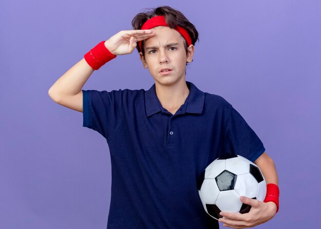 Fronsende jonge knappe sportieve jongen die hoofdband en polsbandjes met beugels draagt die voetbal houden die aan de voorkant kijken en de hand op het voorhoofd houden geïsoleerd op paarse muur