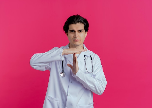 Fronsende jonge mannelijke arts die medische mantel en stethoscoop draagt die time-outgebaar doet dat op roze muur met exemplaarruimte wordt geïsoleerd