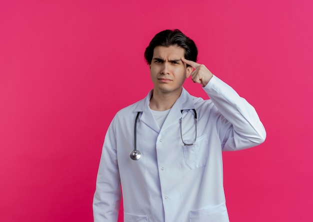 Fronsende jonge mannelijke arts die medische mantel en stethoscoop draagt die vinger op tempel richt die op roze muur met exemplaarruimte wordt geïsoleerd