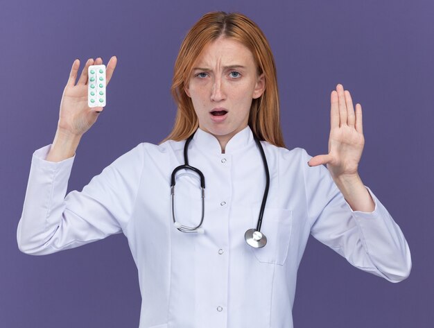 Fronsende jonge vrouwelijke gemberdokter die medische robe en stethoscoop draagt die pakje medische pillen toont aan camera die vijf met hand toont die op purpere muur wordt geïsoleerd