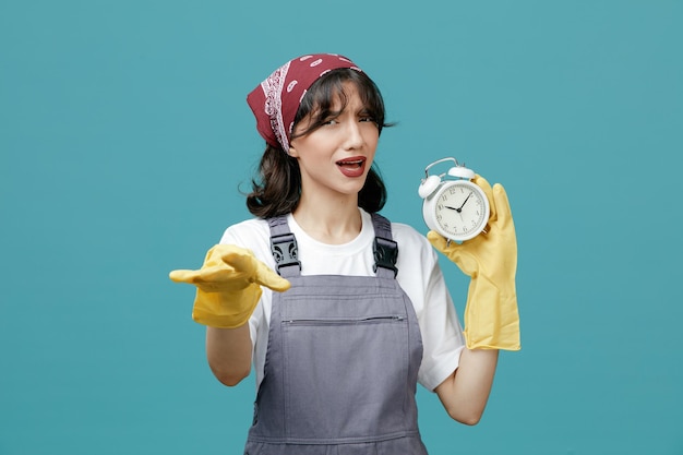 Fronsende jonge vrouwelijke schoonmaakster met uniforme bandana en rubberen handschoenen met wekker die naar camera kijkt en weggaat gebaar geïsoleerd op blauwe achtergrond