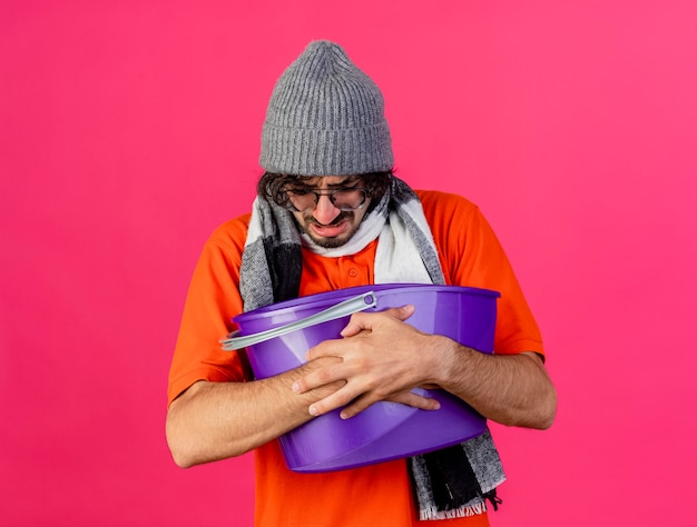 Fronsende jonge zieke man met bril, muts en sjaal met plastic emmer met misselijkheid naar beneden te kijken geïsoleerd op roze muur