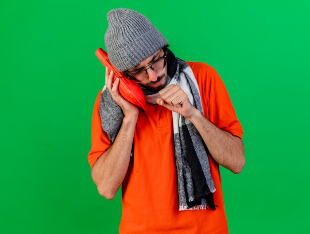 Fronsende jonge zieke man met bril, muts en sjaal warm waterzak op gezicht zetten hoesten vuist houden in de buurt van mond geïsoleerd op groene muur