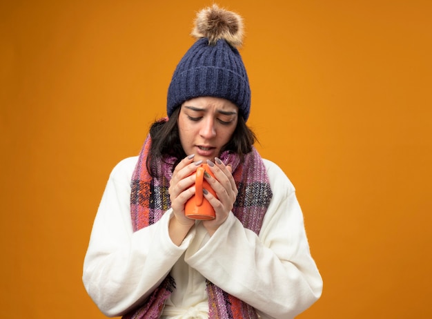 Fronsende jonge zieke vrouw die de muts en de sjaal van de robewinter draagt ?? en binnen kopje thee kijkt die op oranje muur wordt geïsoleerd