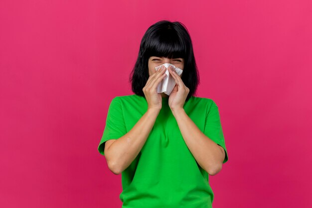 Fronsende jonge zieke vrouw die voorzijde afvegende neus met servet bekijkt dat op roze muur wordt geïsoleerd