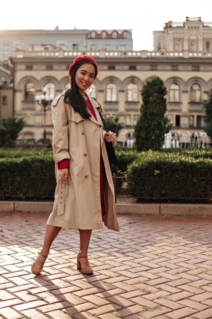 Full-length portret van brunette vrouw in beige trenchcoat, rode baret en jurk glimlacht breed en houdt handtas buiten