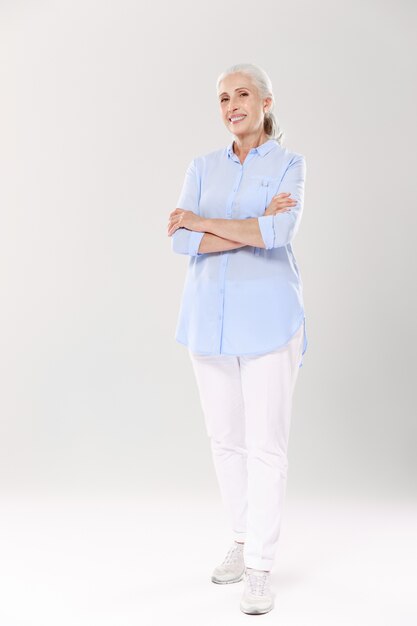 Full-length portret van charmante oude dame in blauw shirt en witte broek, staande met gekruiste handen