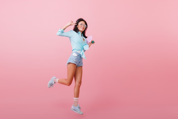 Full-length portret van geïnspireerde donkerharige vrouw in blauw shirt springen en lachen. Blij Spaans meisje met longboard dansen met vredesteken.