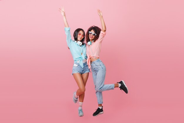 Full-length portret van slank Afrikaans meisje in zwarte sneakers poseren op één been met Europese brunette vriend. Blanke jonge vrouw in blauw shirt grappig dansen met mulat dame.