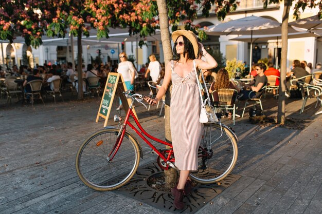 Full-length portret van trendy meisje in hoed en lange jurk staande op terras met fiets