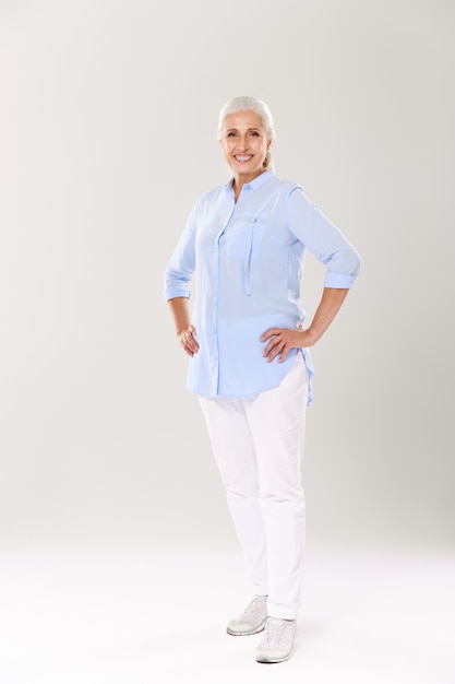 Full-length portret van vrolijke volwassen vrouw in blauw shirt en witte broek, permanent met handen op haar middel