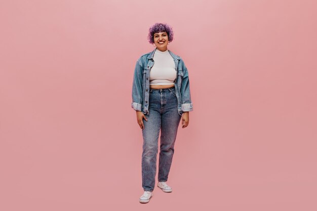 Full-length portret van vrolijke vrouw met kort paars haar in denim pak, witte sneakers en lichte top glimlachen