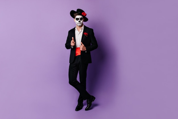 Full-length shot van charmante man met rode satijnen riem onder zwarte jas, poseren in Halloween kostuum.