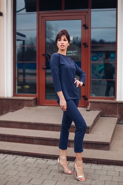 Full length stock fashion portret van een mooie brunette in blauw sportief pak met trui en hoge hakken poseren in de straat voor het gebouw. Sportchique stijl. Mode-concept.