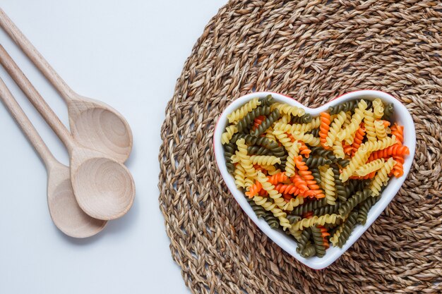 Fusilli pasta in een hartvormige kom met houten lepels bovenaanzicht op witte en rieten placemat tafel