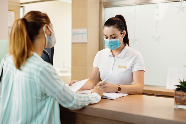 Fysiotherapeut met gezichtsmasker die gegevens van haar cliënt schrijft bij receptie